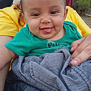 Lucille is registered to the contest to win money with this photo: baby, child, tongue_out, green_shirt, yellow_shirt, blanket, hand, outdoor, nature, person, smile, portrait, cute, infant, face, young, happy, clothing, tree, holding