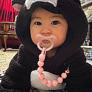 Demeri is registered to the contest to win money with this photo: baby, child, pacifier, costume, bear_costume, hood, indoors, carpet, furniture, red_curtain, living_room, cute, adorable, pink_beads, face, person, childwear, home, cozy, floor