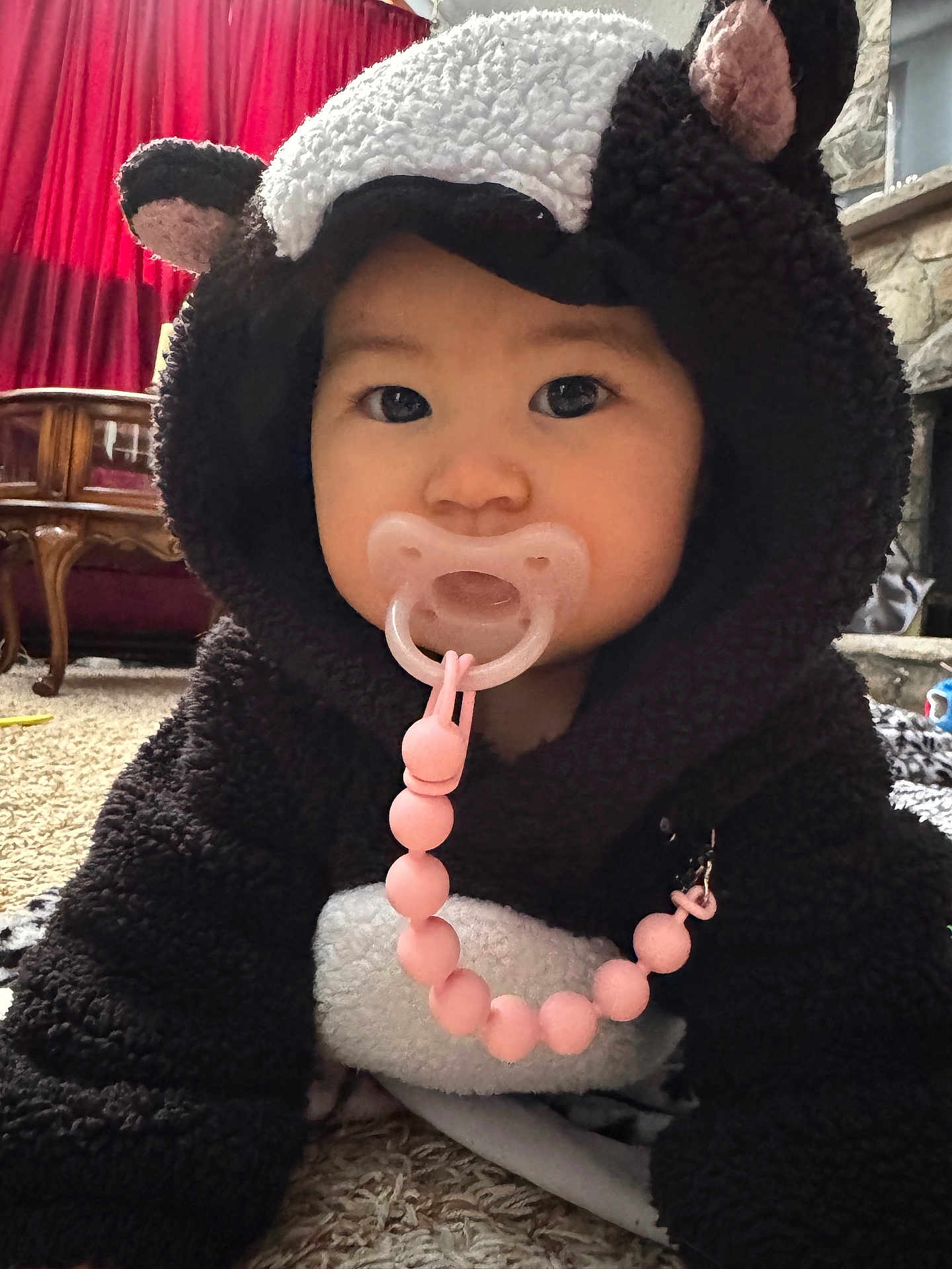 Demeri is registered to the contest to win money with this photo: baby, child, pacifier, costume, bear_costume, hood, indoors, carpet, furniture, red_curtain, living_room, cute, adorable, pink_beads, face, person, childwear, home, cozy, floor