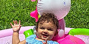 Oaklen joined the competition — help win amazing prizes! toddler, child, water, pool, inflatable, pink, green, grass, outdoor, summer, splashing, happy, smiling, curly_hair, blue_clothing, sunlight, playful, toy, water_droplets, fence