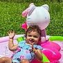 toddler, child, water, pool, inflatable, pink, green, grass, outdoor, summer, splashing, happy, smiling, curly_hair, blue_clothing, sunlight, playful, toy, water_droplets, fence
