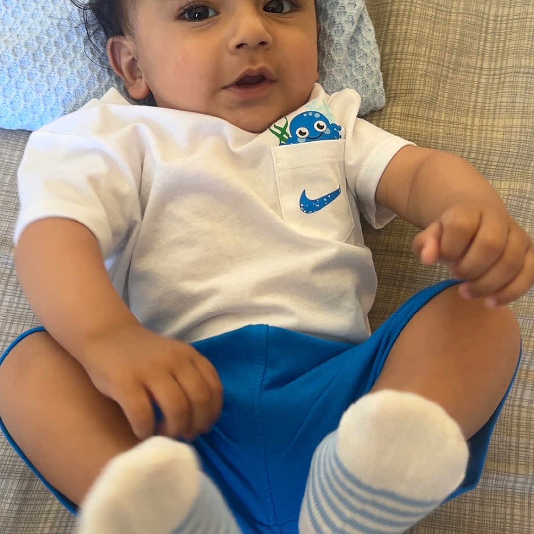 Nirvaan is registered to the contest to win money with this photo: baby, bed, blue_shorts, child, clothing, curious, cute, eyes, face, footwear, hair, hand, indoors, infant, lying_down, pillow, playful, portrait, socks, white_shirt