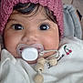 baby, pacifier, pink_hat, white_outfit, close_up, face, big_eyes, curly_hair, blanket, beads, cute, infant, child, indoors, soft_texture, warm_clothing, portrait, smiling_eyes, cozy, toddler