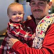 Sylvia is registered to the contest to win money with this photo: baby, child, man, adult, holding, plaid, red_clothing, cap, glasses, blanket, indoor, portrait, smile, face, person, happy, casual, beard, fabric, warm_clothing