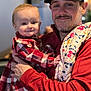 baby, child, man, adult, holding, plaid, red_clothing, cap, glasses, blanket, indoor, portrait, smile, face, person, happy, casual, beard, fabric, warm_clothing