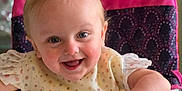 Sylvia is registered to the contest to win money with this photo: baby, child, high_chair, smiling, happy, polka_dot, bib, face, arm, furniture, indoor, person, hair, table, seat, pink, white, cute, expression, playful