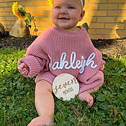 Oakleigh is registered to the contest to win money with this photo: baby, child, infant, pink_sweater, white_bow, grass, outdoor, sunlight, flower, yellow_flower, brick_wall, smiling, happy, cute, seven_months, headband, barefoot, portrait, nature, sitting