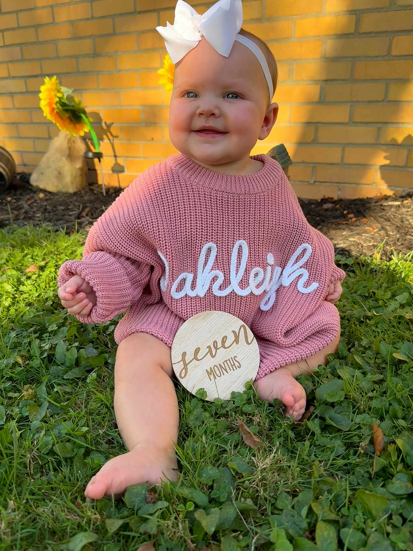 Oakleigh is registered to the contest to win money with this photo: baby, child, infant, pink_sweater, white_bow, grass, outdoor, sunlight, flower, yellow_flower, brick_wall, smiling, happy, cute, seven_months, headband, barefoot, portrait, nature, sitting