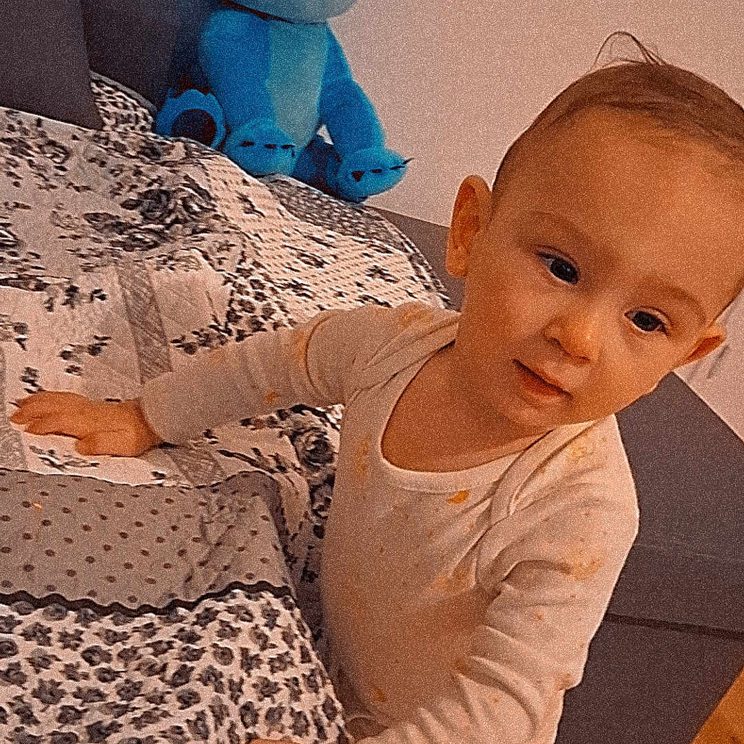 Mathias Nicolas participe au concours pour gagner de l'argent avec cette photo : bed, blue, carpet, child, couch, curious, ear, face, hand, home, indoor, leaning, pajamas, patterned_bedspread, plush_toy, smiling, stitch, toddler, toy, wood_floor