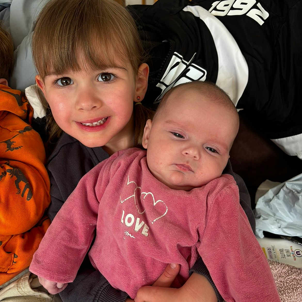 Judith a rejoint le concours — aidez-le/la à gagner de superbes lots ! child, baby, smiling, holding, pink_clothing, girl, infant, indoor, family, love, cute, portrait, casual, happy, sibling, person, face, head, cuddling, home