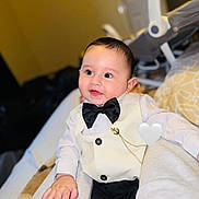 Thiago is registered to the contest to win money with this photo: accessory, baby, bed, black_pants, blanket, bow_tie, chain, child, cute, face, formal_clothing, hand, happy, indoors, infant, person, seated, smile, toddler, vest