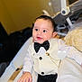 accessory, baby, bed, black_pants, blanket, bow_tie, chain, child, cute, face, formal_clothing, hand, happy, indoors, infant, person, seated, smile, toddler, vest