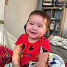 baby, child, infant, smiling, portrait, indoors, bedroom, bed, blanket, red_outfit, cartoon_clothing, sitting, happy, face, person, television, shelving, home, fuzzy_blanket, closeup
