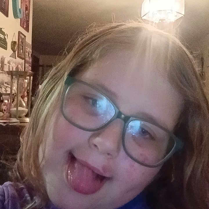 Isabelle is registered to the contest to win money with this photo: child, glasses, tongue_out, indoor, light_fixture, wall_art, purple_clothing, smiling, face, person, home, cozy, portrait, hair, casual, funny_face, young, playful, room, decor