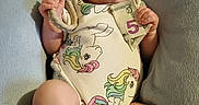 Iris joined the competition — help win amazing prizes! baby, bear, child, clothing, cushion, cute, face, five_dollar_bill, foot, hand, indoor, infant, money, onesie, pacifier, person, portrait, relaxing, toy, unicorn