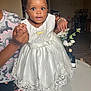 baby, child, dress, white_dress, lace, hair_accessories, bracelet, necklace, indoor, living_room, adult_hands, flower_vase, table, furniture, portrait, cute, smiling, person, holding, happy