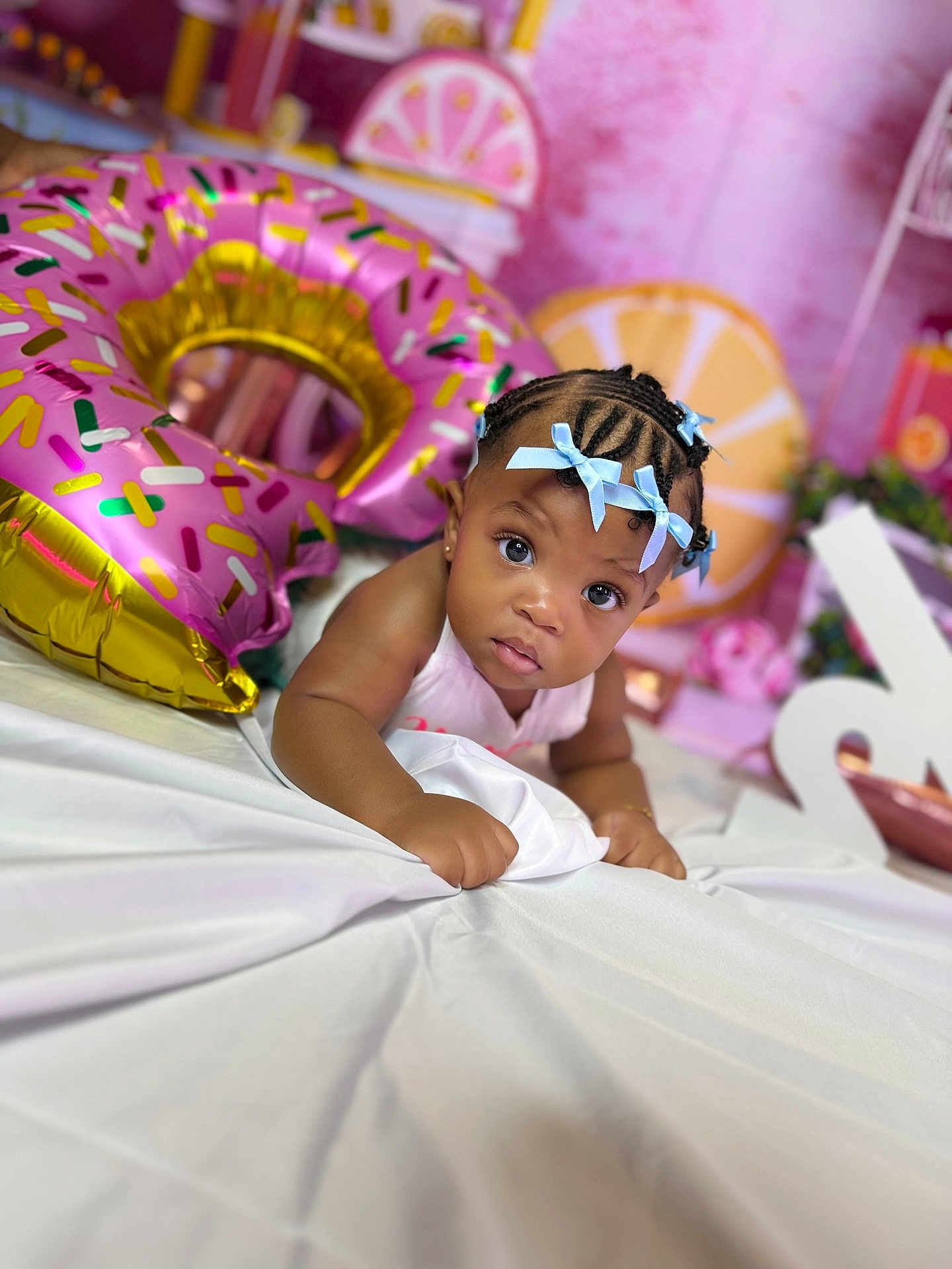 Kaira is registered to the contest to win money with this photo: baby, child, braids, blue_bows, pink_balloon, donut_balloon, white_sheet, indoor, curious, decorations, celebration, portrait, infant, face, cute, person, closeup, colorful, festive, smiling