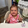 Vera is registered to the contest to win money with this photo: baby, child, high_chair, smile, pink_clothing, pacifier, kitchen, stove, towel, happy, indoor, face, person, infant, cute, expression, sitting, toy, hair_tie, table