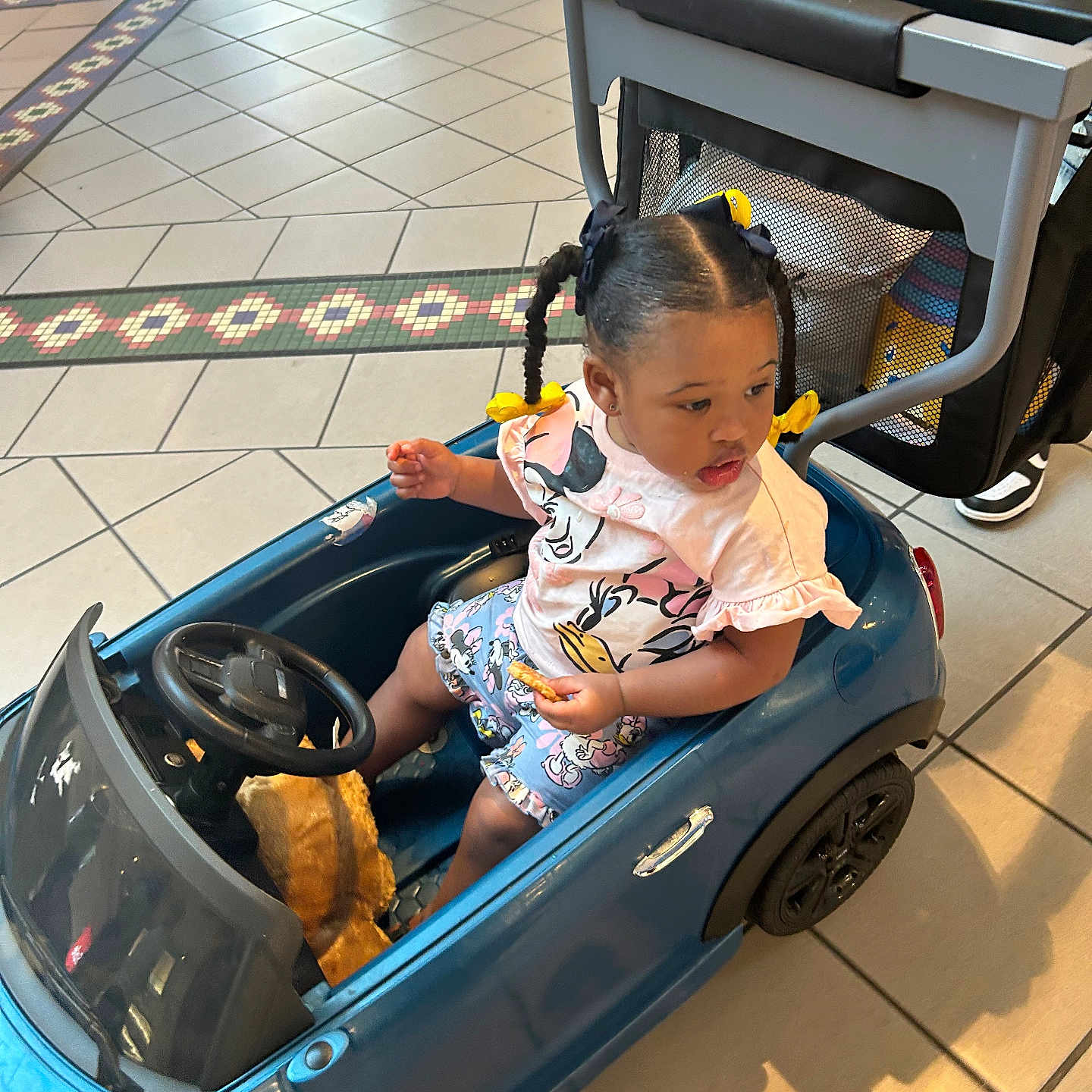 Keoir is registered to the contest to win money with this photo: braided_hair, casual_clothing, child, face, floor_tile, footwear, hand, handle, indoor, person, seat, shorts, snack, stroller, toddler, toy_car, tshirt, wheel, window, yellow_bows