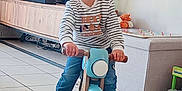 Noah participe au concours pour gagner de l'argent avec cette photo : toddler, child, balance_bike, indoor, striped_shirt, smiling, toy, living_room, floor_tiles, wooden_furniture, plush_toy, game_board, casual_clothing, sneakers, happy, person, home, play, cute, young