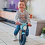 toddler, child, balance_bike, indoor, striped_shirt, smiling, toy, living_room, floor_tiles, wooden_furniture, plush_toy, game_board, casual_clothing, sneakers, happy, person, home, play, cute, young