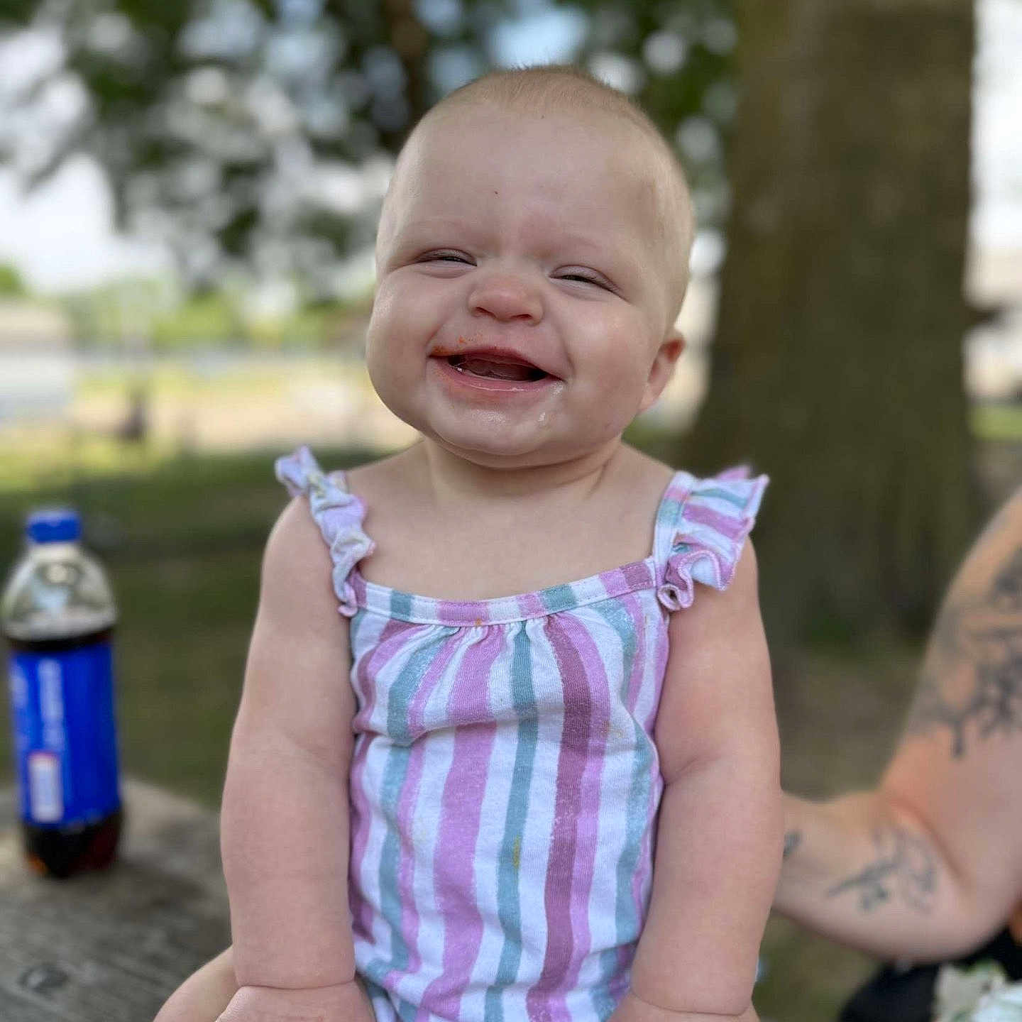 Roselie is registered to the contest to win money with this photo: arm, baby, bottle, child, cute, drink, face, happy, messy, nature, outdoor, picnic_table, playful, sitting, smiling, striped_clothing, summer, tattoo, tree, wood