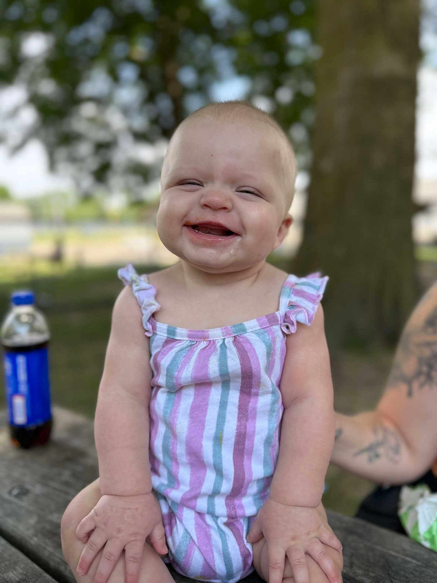 Roselie is registered to the contest to win money with this photo: baby, child, smiling, outdoor, picnic_table, striped_clothing, happy, face, arm, tattoo, bottle, drink, summer, nature, tree, wood, sitting, cute, messy, playful
