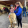 child, pony, helmet, woman, animal, outdoor, riding, horse, person, barn, shelter, casual_clothing, gravel_ground, daylight, support, holding, leisure, fun, smile, pet