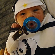 Kaspian is registered to the contest to win money with this photo: baby, pacifier, hat, plush_toy, giraffe, panda, clothing, infant, face, cute, indoors, child, toy, blanket, sleepwear, cozy, young, portrait, lying_down, comfort