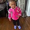 child, toddler, girl, portrait, standing, pink_jacket, varsity_jacket, purple_shoes, crocs, hair_bows, earrings, looking_down, indoors, wooden_floor, stroller, sofa, lamp, home, clothing, smiling