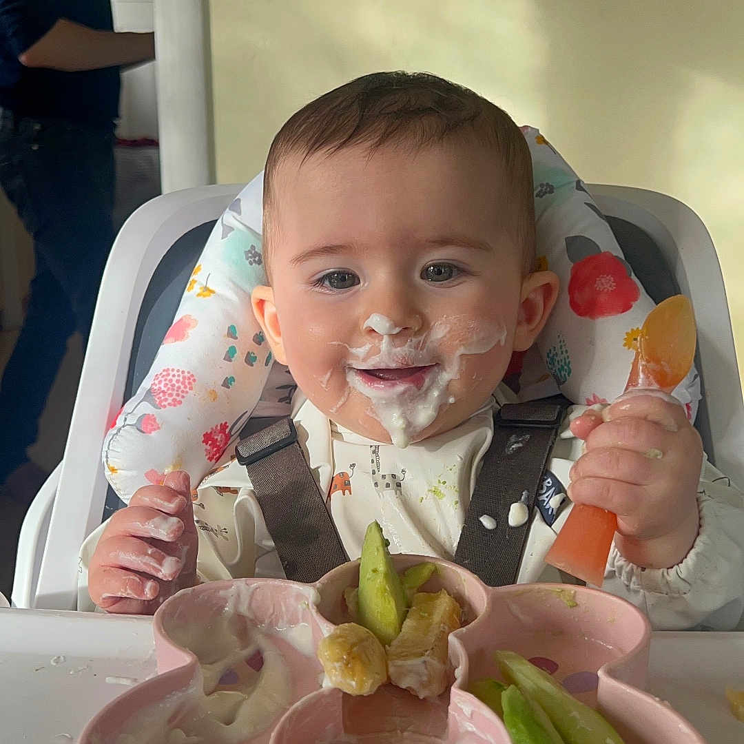 Nina joined the competition — help win amazing prizes! adult, baby, background, bib, child, clothing, feeding, food, furniture, happy, high_chair, home, indoor, light_switch, messy, person, plate, smiling, spoon, yogurt