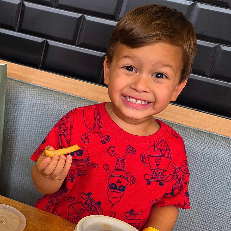 Addiel joined the competition — help win amazing prizes! child, boy, smiling, grinning, restaurant, booth, food, french_fries, snack, plate, table, red_shirt, cartoon_shirt, seated, portrait, indoor, dining, happy, face, meal