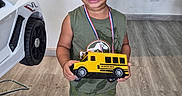 Addiel is registered to the contest to win money with this photo: child, boy, toddler, smiling, medal, toy, school_bus, yellow, indoor, portrait, standing, playful, shorts, sandals, green_clothing, wood_floor, toy_car, vehicle, home, happy