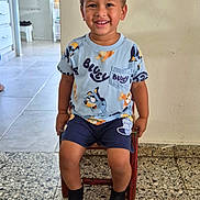 Addiel is registered to the contest to win money with this photo: child, boy, toddler, smiling, portrait, sitting, chair, indoor, home, hallway, tile_floor, pajamas, bluey, cartoon, shorts, socks, brown_hair, happy, playful, family_photo