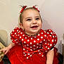 child, toddler, smile, red_dress, polka_dots, bow, pigtails, happy, portrait, indoor, white_wall, necklace, cheeks, eyes, teeth, sitting, hand, tulle_skirt, fabric, cute