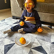 Myles joined the competition — help win amazing prizes! toddler, child, sunglasses, orange, fruit, bowl, rug, carpet, couch, cushion, indoor, living_room, sitting, smiling, happy, curly_hair, clothing, sock, person, home