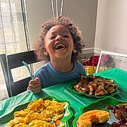 Myles is registered to the contest to win money with this photo: child, smiling, curly_hair, blue_shirt, fork, table, green_tablecloth, scrambled_eggs, bread, donut, grapes, plate, food, indoor, window, chair, happy, meal, breakfast, person