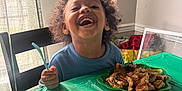 Myles is registered to the contest to win money with this photo: child, smiling, curly_hair, blue_shirt, fork, table, green_tablecloth, scrambled_eggs, bread, donut, grapes, plate, food, indoor, window, chair, happy, meal, breakfast, person