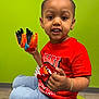 toddler, child, red_shirt, jeans, toy, indoor, green_wall, television, cushion, sitting, smiling, playful, bracelet, floor, casual_clothing, portrait, person, young_child, curious, bright_colors