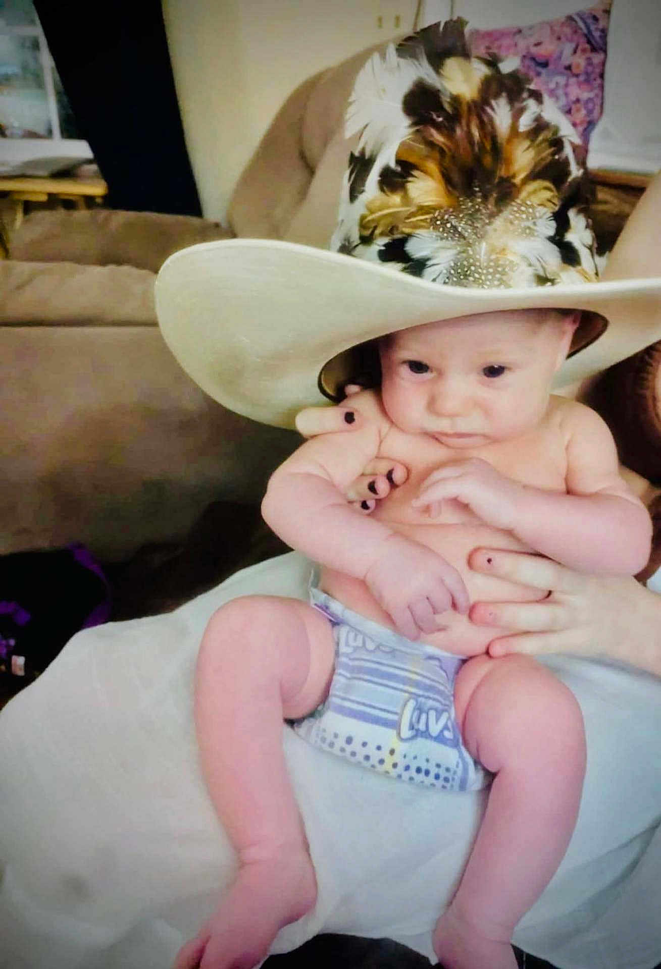 Keegan joined the competition — help win amazing prizes! baby, infant, hat, cowboy_hat, feathers, diaper, person, hand, nail_polish, furniture, couch, living_room, skin, legs, arms, cute, portrait, indoor, holding, seated