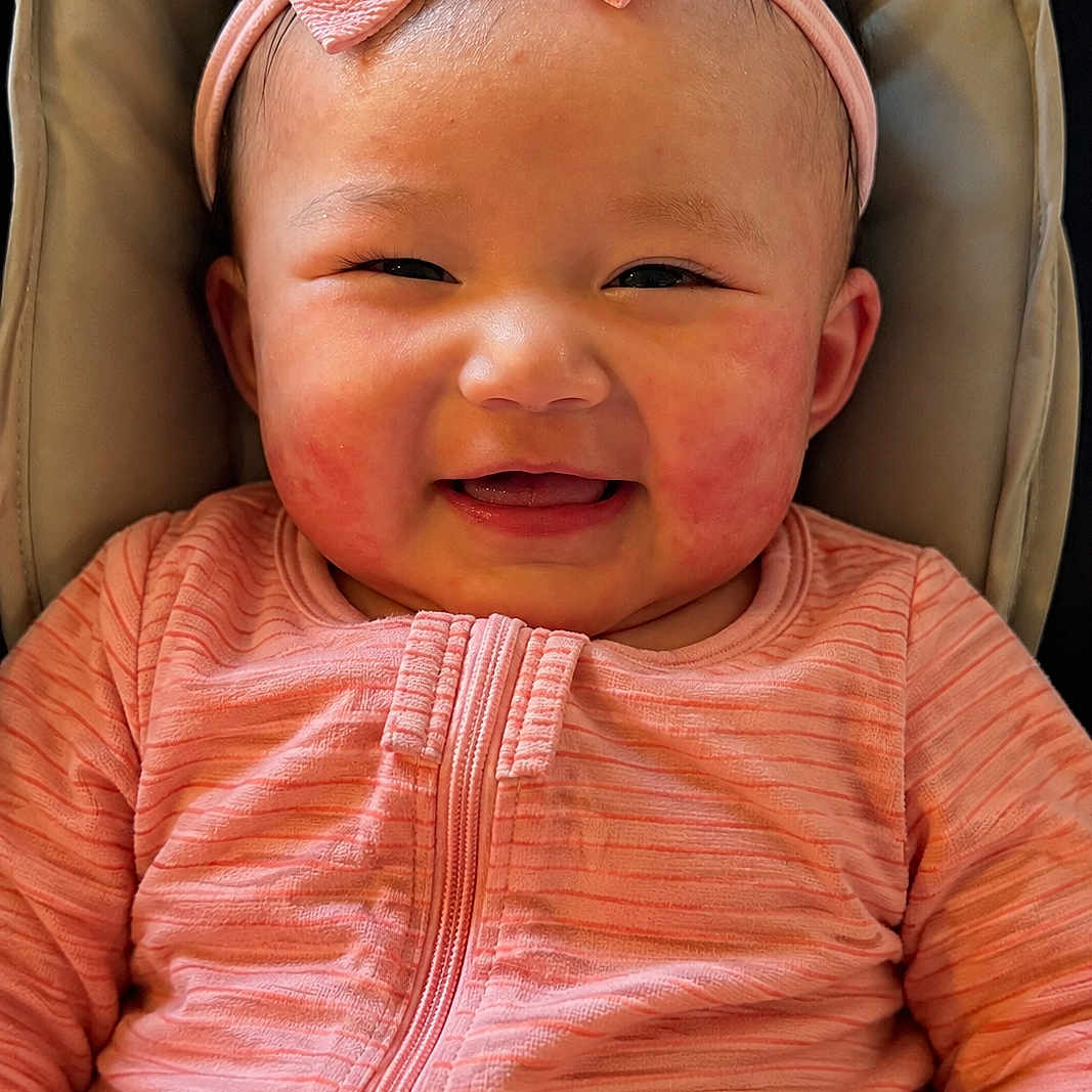 Sofia is registered to the contest to win money with this photo: baby, infant, smiling, pink_clothing, headband, bow, face, cheerful, cushion, seated, portrait, child, happy, indoors, closeup, person, cute, young_child, apparel, zipped_jacket