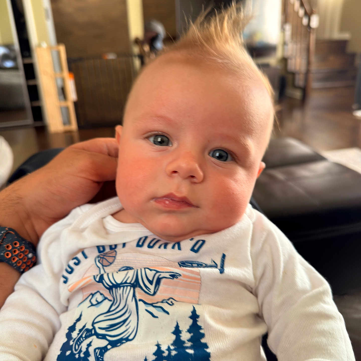 Liam joined the competition — help win amazing prizes! baby, infant, child, portrait, close_up, face, blue_eyes, mohawk, hair, wide_eyed, neutral_expression, white_shirt, graphic_shirt, held, adult_hand, indoor, home, living_room, staircase, couch