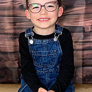 Ethan participe au concours pour gagner de l'argent avec cette photo : child, smiling, glasses, denim_overalls, black_shirt, hands, wooden_floor, wooden_background, portrait, happy, indoor, cute, young, fashion, sitting, eyewear, person, casual, kid, studio