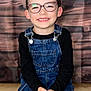 child, smiling, glasses, denim_overalls, black_shirt, hands, wooden_floor, wooden_background, portrait, happy, indoor, cute, young, fashion, sitting, eyewear, person, casual, kid, studio