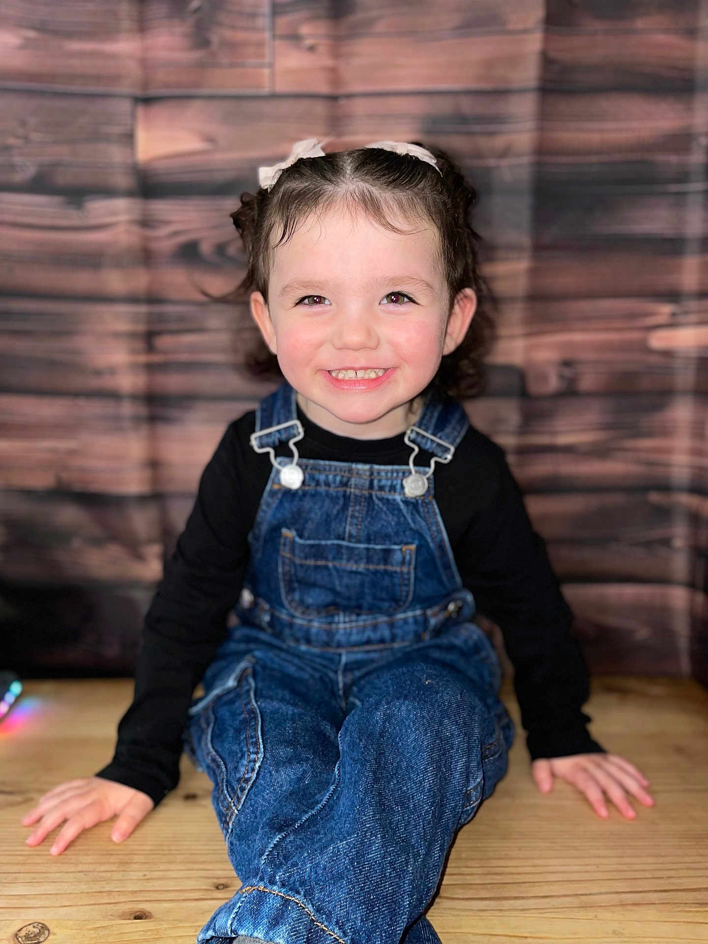 Luna participe au concours pour gagner de l'argent avec cette photo : toddler, child, smiling, denim_overalls, black_shirt, curly_hair, sitting, wooden_floor, wooden_background, portrait, happy, face, person, cute, indoors, young_child, casual_clothing, looking_at_camera, bright_eyes, playful