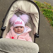Delilah is registered to the contest to win money with this photo: baby, baby_carriage, child, cute, daylight, face, happy, hat, infant, jacket, outdoor, person, pink, pom_poms, sidewalk, smile, stroller, transportation, urban, warm_clothing