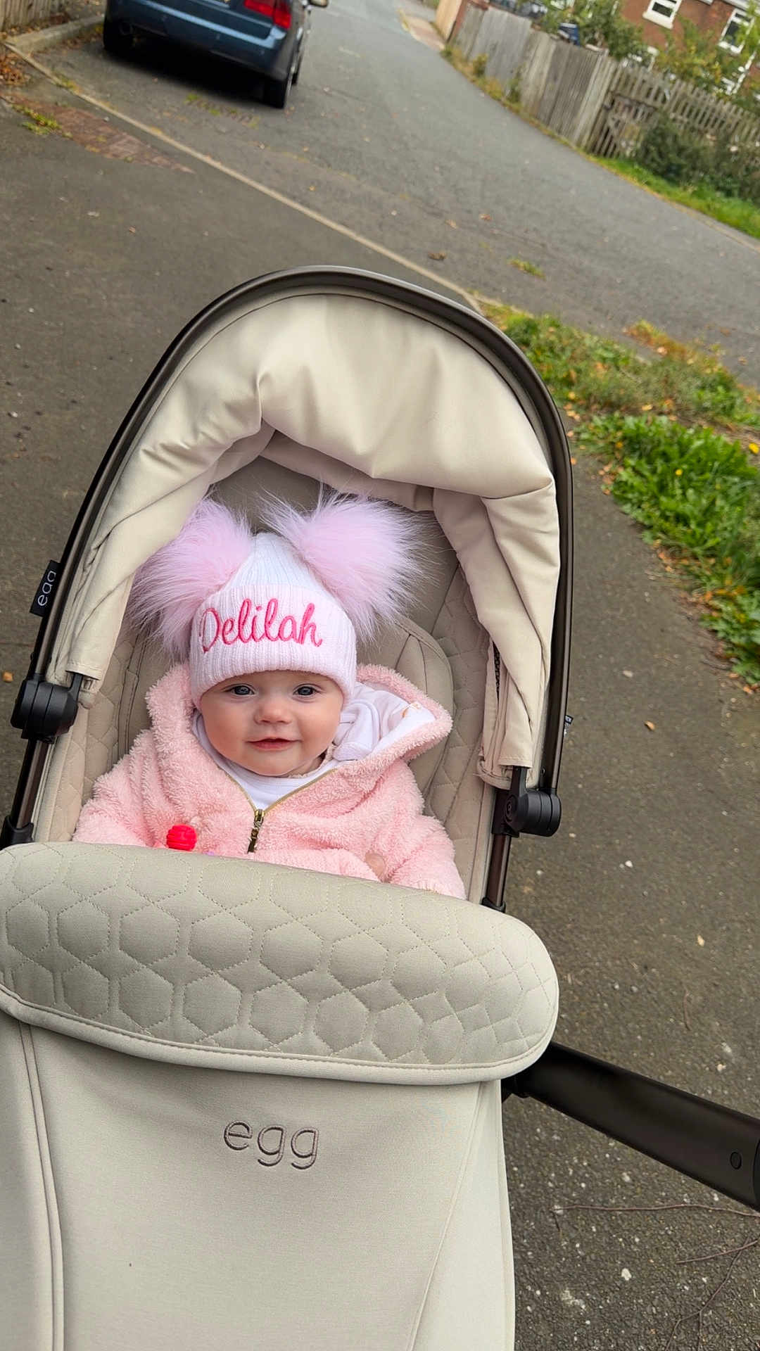 Delilah is registered to the contest to win money with this photo: baby, baby_carriage, child, cute, daylight, face, happy, hat, infant, jacket, outdoor, person, pink, pom_poms, sidewalk, smile, stroller, transportation, urban, warm_clothing