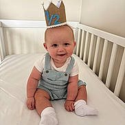 Rowan is registered to the contest to win money with this photo: baby, crown, crib, smile, overalls, white_shirt, socks, celebration, milestone, indoor, child, cute, happy, infant, sitting, face, person, headwear, white_background, soft_lighting