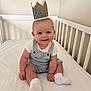baby, crown, crib, smile, overalls, white_shirt, socks, celebration, milestone, indoor, child, cute, happy, infant, sitting, face, person, headwear, white_background, soft_lighting