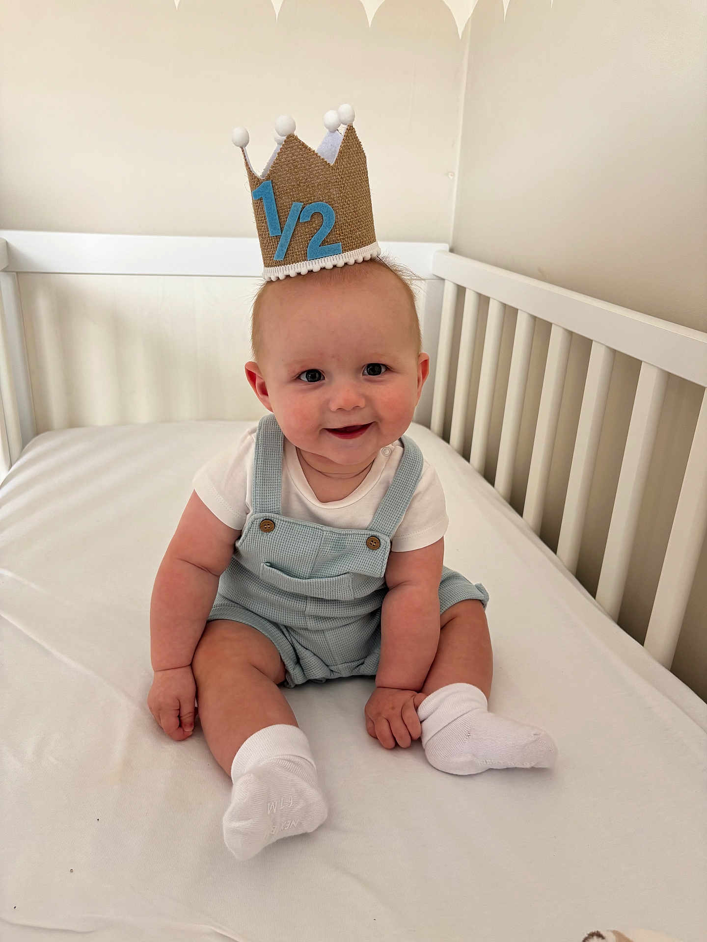 Rowan is registered to the contest to win money with this photo: baby, crown, crib, smile, overalls, white_shirt, socks, celebration, milestone, indoor, child, cute, happy, infant, sitting, face, person, headwear, white_background, soft_lighting